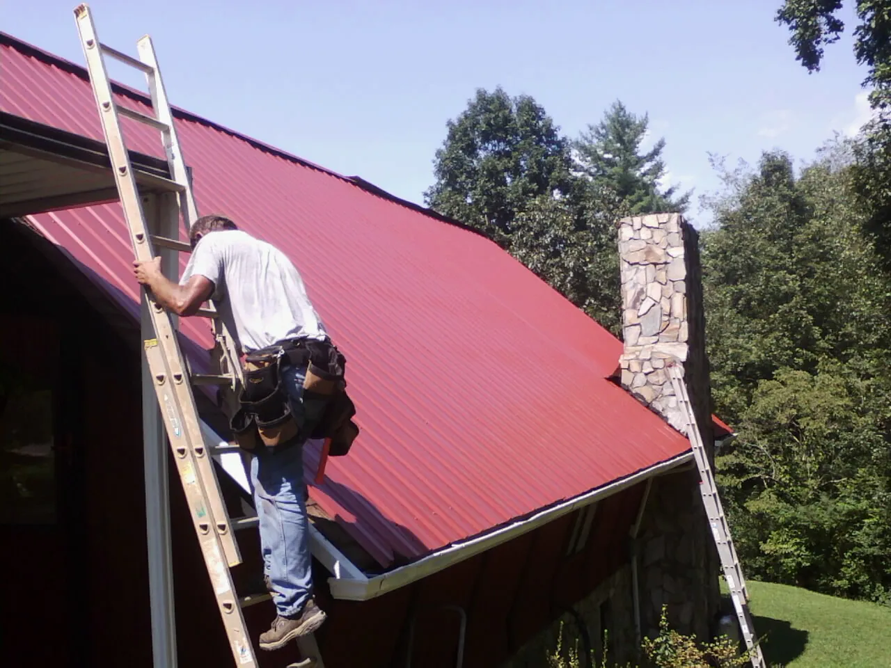 Roofer on a ladder beside a red metal roof and stone chimney — Soffit & Fascia Repair by Roofing Brothers in Dry Branch